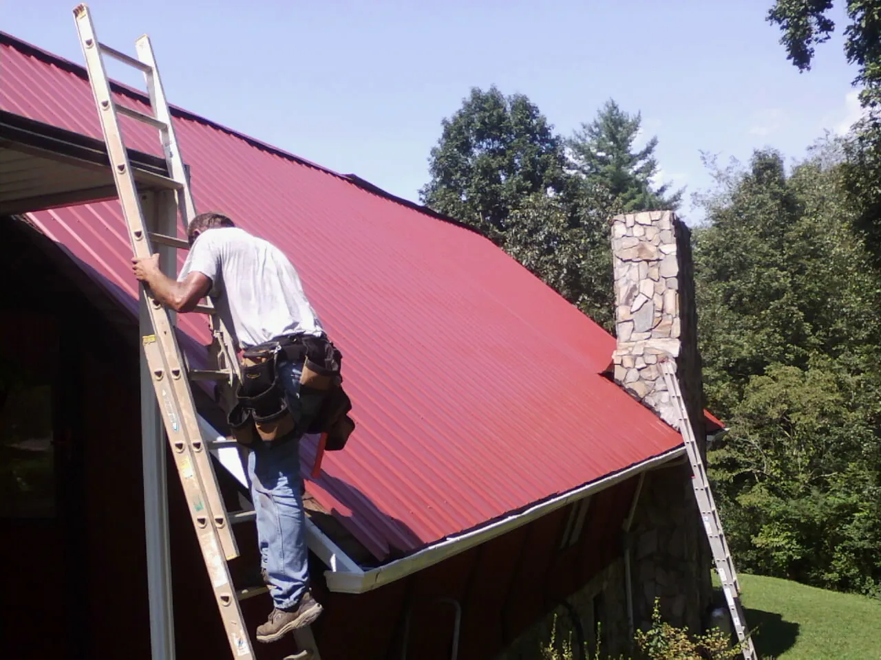 Roofer on a ladder beside a red metal roof and stone chimney — Soffit & Fascia Repair by Roofing Brothers in Mont Vernon